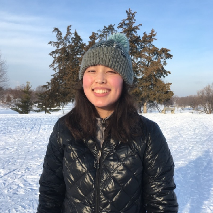 woman with dark hair, a gray beanie, and black jacket stands smiling in front of a snowy field