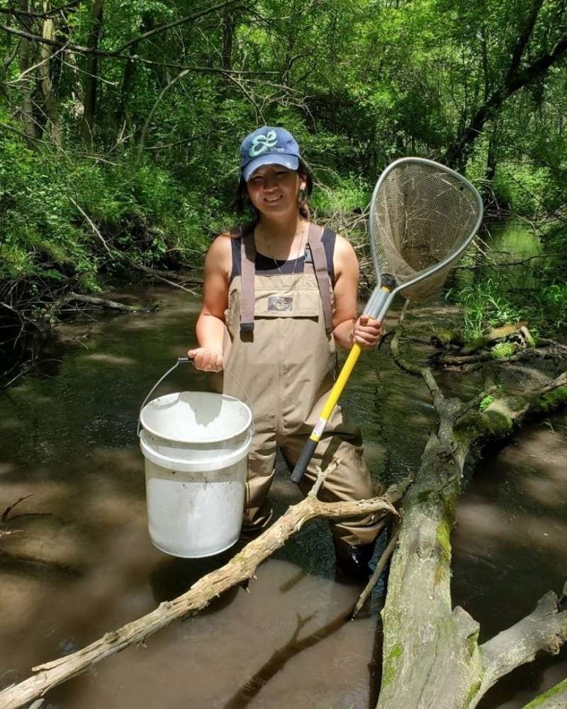 a woman in a hat stands in a creek with a white bucket and fishing net