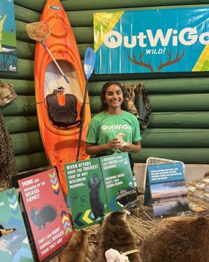 a young woman stands behind a table with pamphlets and a kayak in the background