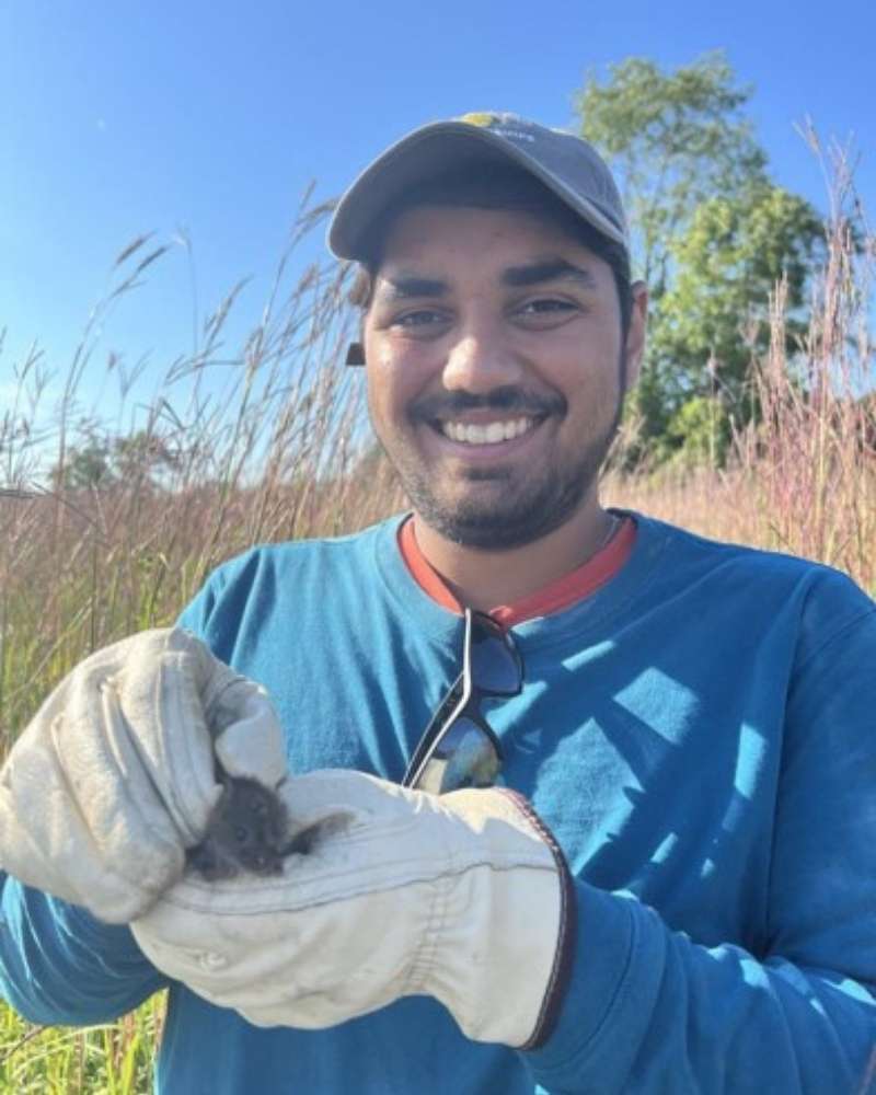 a man in a hat stands in a prairie holding a small mammal in gloved hands