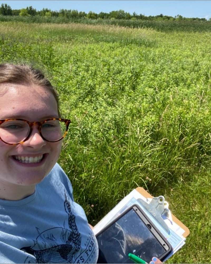 young woman stands in a prairie with a clipboard and tablet
