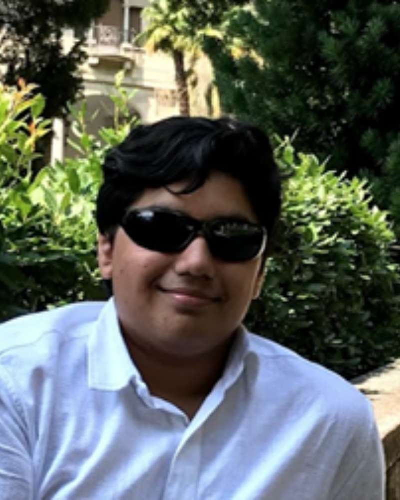 man with dark hair, sunglasses, and a white shirt stands in front of green landscaping