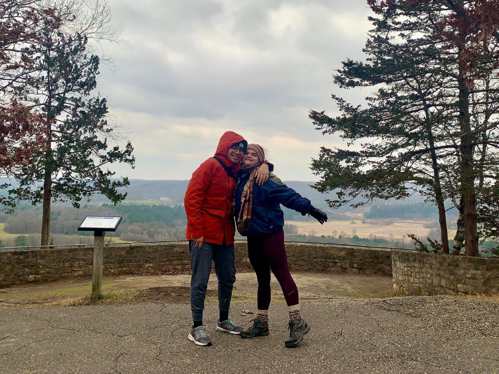 Two people in warm clothes in front of a lookout point at a state park, surrounded by tall trees overlooking a cloudy sky and green bluffs