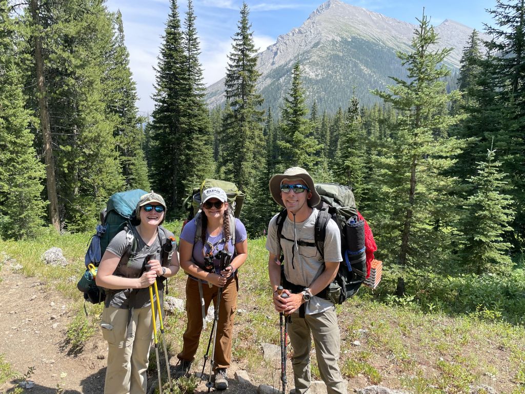 Emma and two of her friends with backpacking gear on in front of a mountain and pine trees
