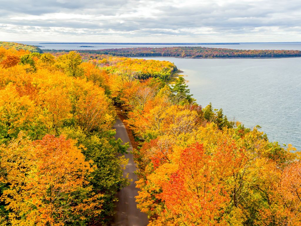 Bird’s eye view of a road with bright, colorful trees on either side with a lake and small islands in the background..