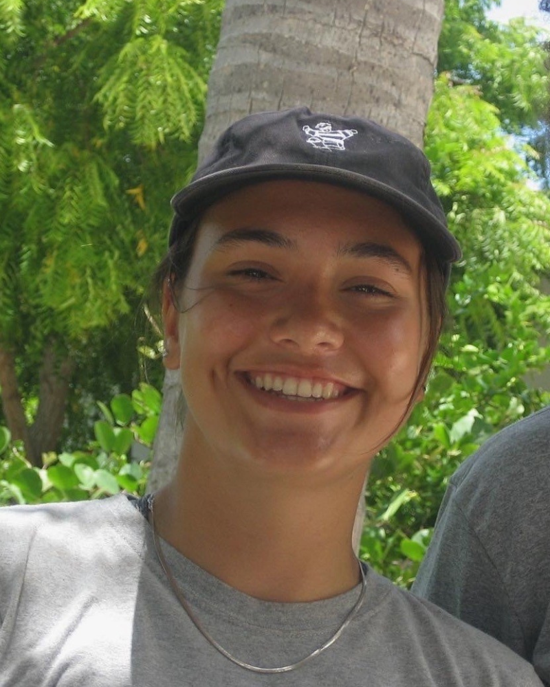 Raquel woman with dark hair, a gray beanie, and black jacket stands smiling in front of a snowy field