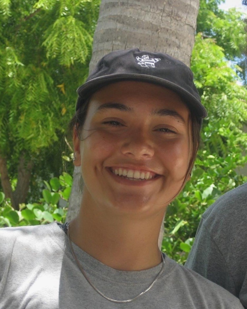 woman with dark hair, a gray beanie, and black jacket stands smiling in front of a snowy field