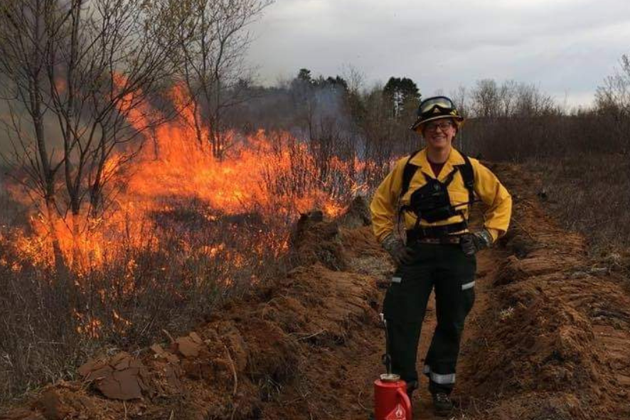 Photo description: Carly Lapin, in fire gear, standing next to a prescribed burn at Spread Eagle State Natural Area. 