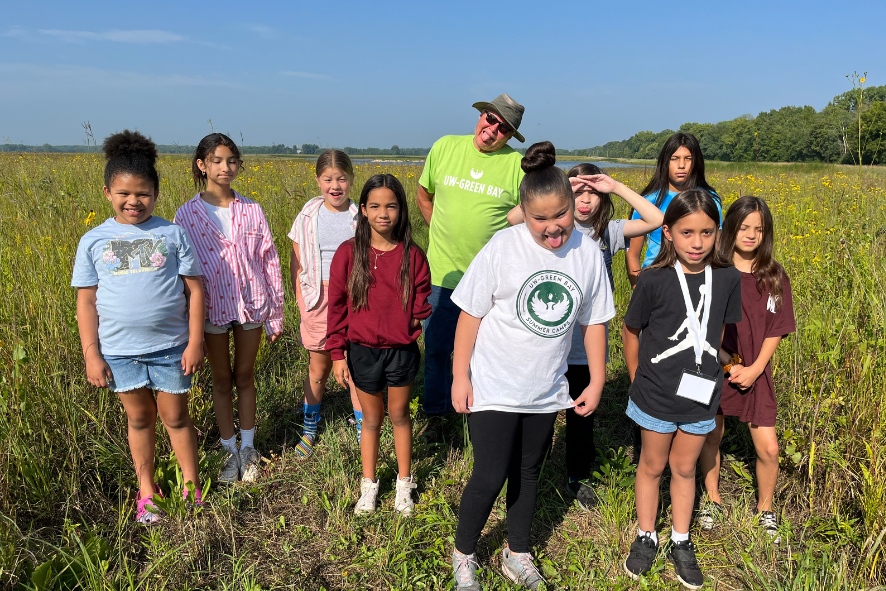 Students and staff making silly faces while standing in a prairie during a field trip during the Oneida summer camp program