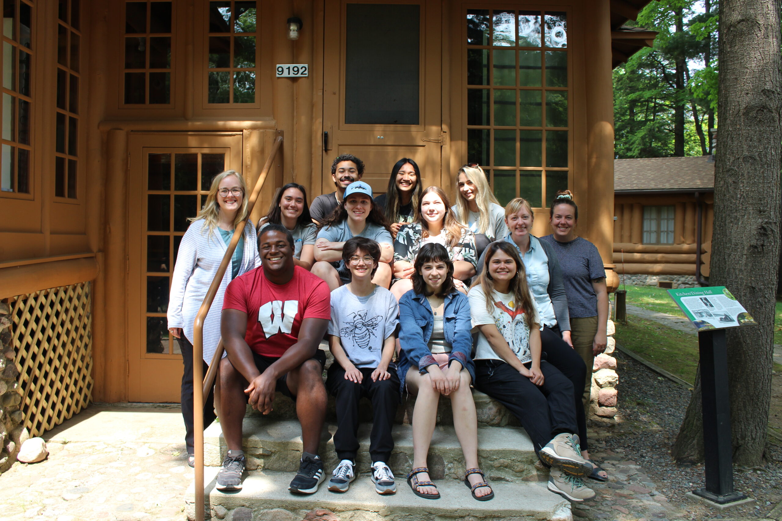 Students sitting on the steps in front of a building with two adults standing to the left and right of the group