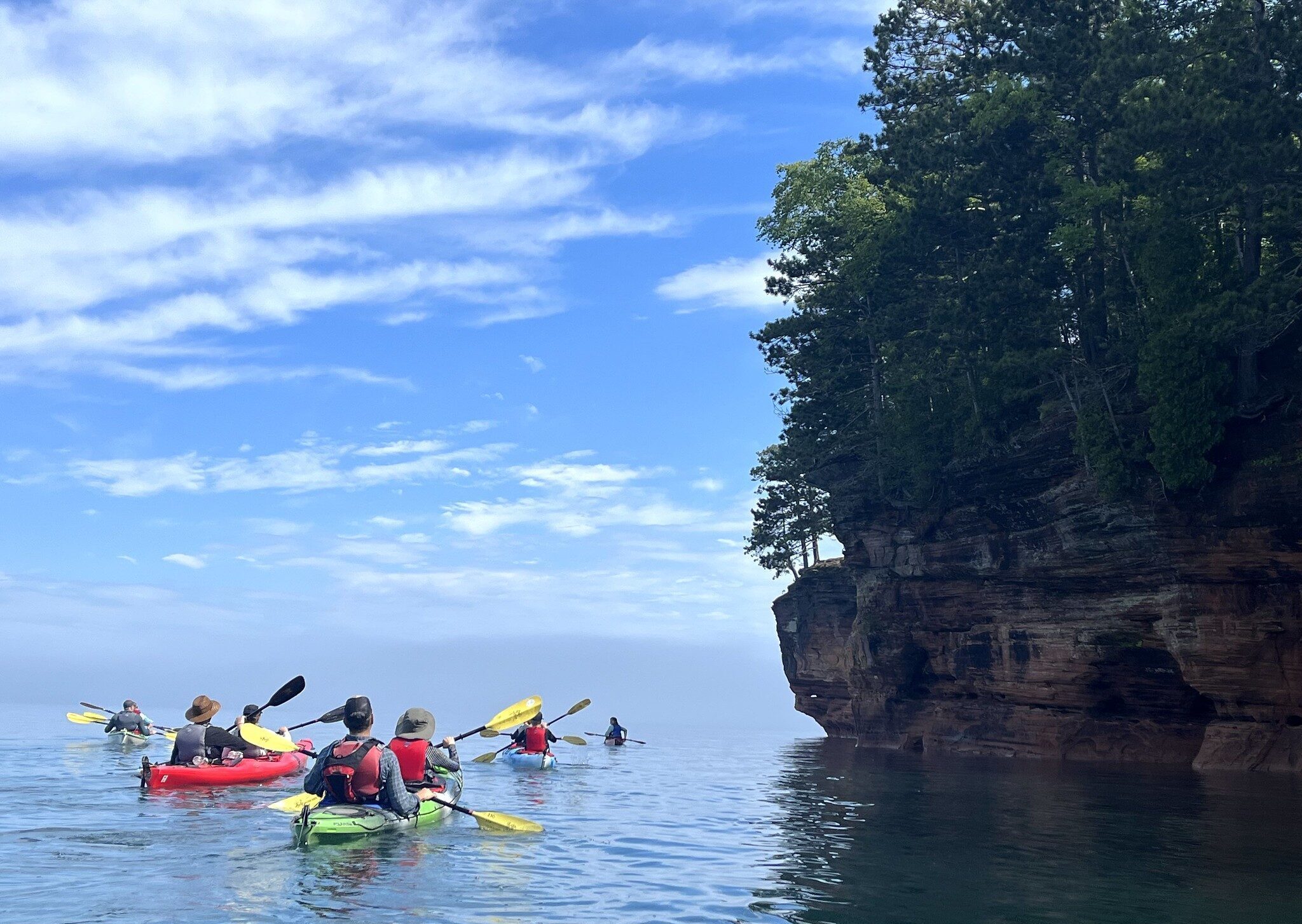 People kayaking on the water next to a piece of land and tall trees sticking out across the shore